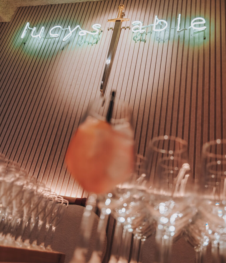 Neonlogo van Lucy’s Table boven de bar met wijnglazen en cocktail in warme verlichting